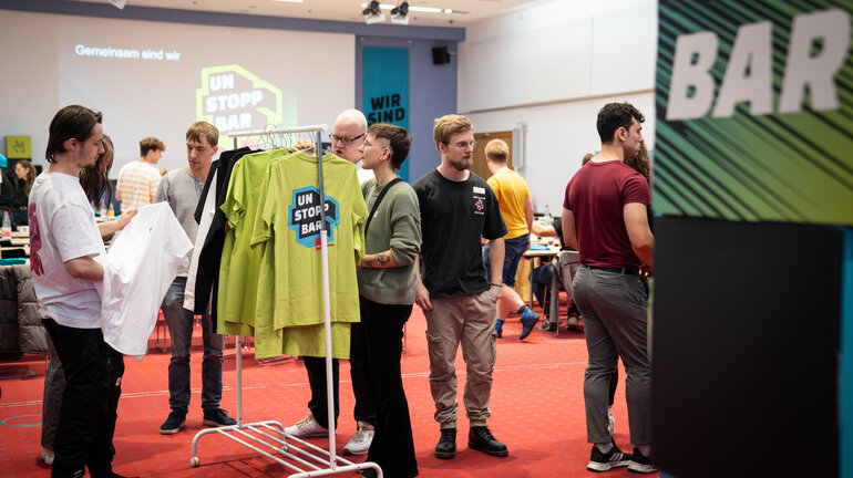 Mehrere Personen stehen an einer Kleiderstange und schauen grüne T-Shirts im Kampagnendesign an.