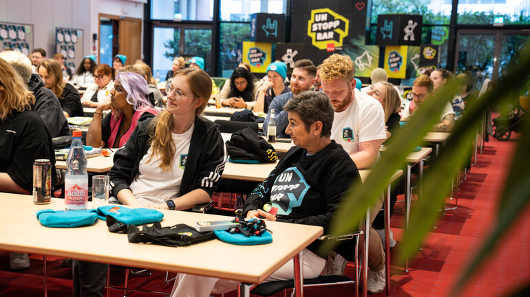 viele Menschen sitzen in einem Konferenzraum und schauen zu einer Bühne. Im Hintergrund sind Würfel mit UNSTOPPBAR-Design.