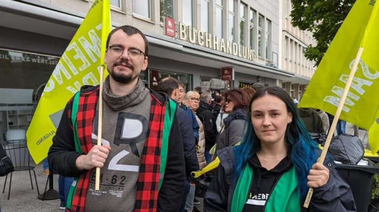 Zwei Personen mit grünen Westen und gelben Fahnen schauen in die Kamera am bundesweiten Jugendaktionstag in der Tarifrunde Telekom 2024