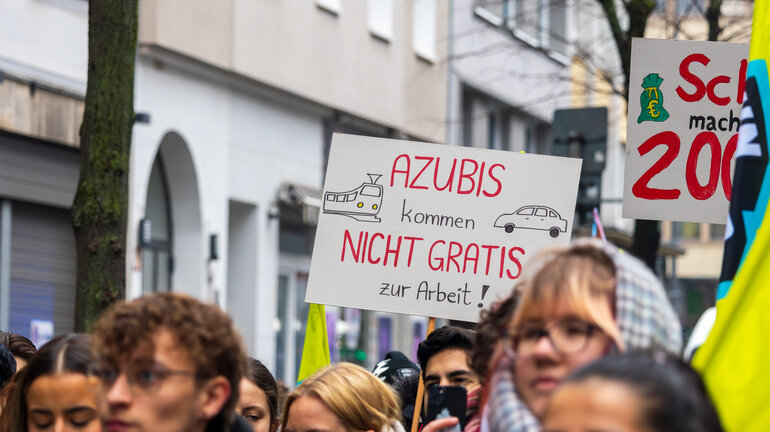 Man sieht Köpfe von streikenden Personen sowie die Schilder, die sie hochhalten. Auf einem steht "Azubis kommen nicht gratis zur Arbeit".