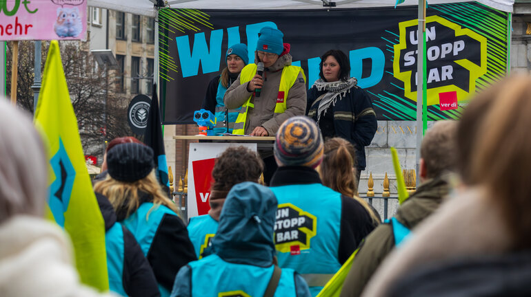 Auf einer Bühne steht eine Person in einer Streikweste vor einem Banner mit der Aufschrift "Wir sind Unstoppbar" und spricht zu einer Gruppe Personen in Streikwesten.