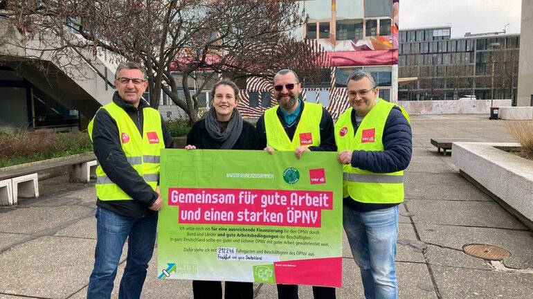 Grüne MdB Deborah Düring posiert mit drei Streikenden und einem Poster mit Forderungen.