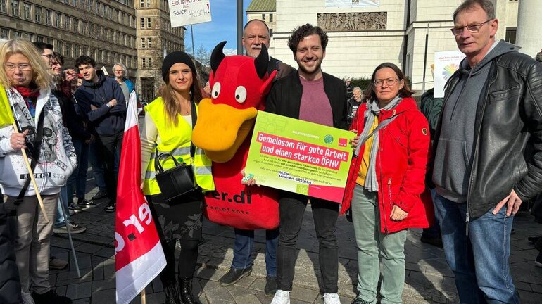 der Grüne MdB Felix Banaszek posiert mit Streikenden und einem Streikposter mit Forderungen.