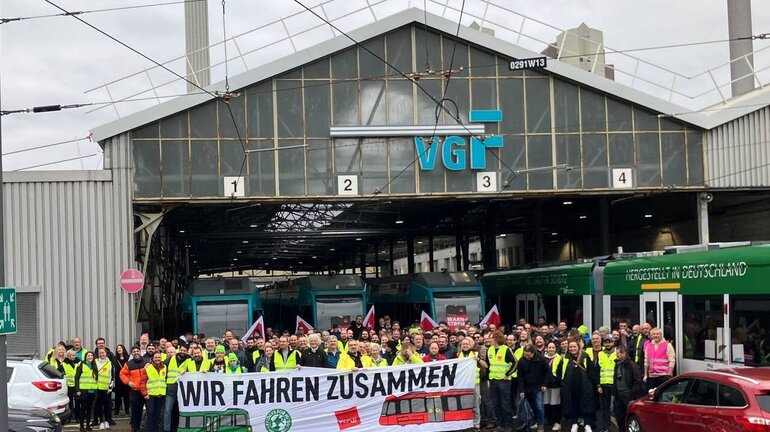 Eine große Gruppe Streikender steht vor einem Trambahnhof mit einem Banner mit der Aufschrift Wir Fahren Zusammen.