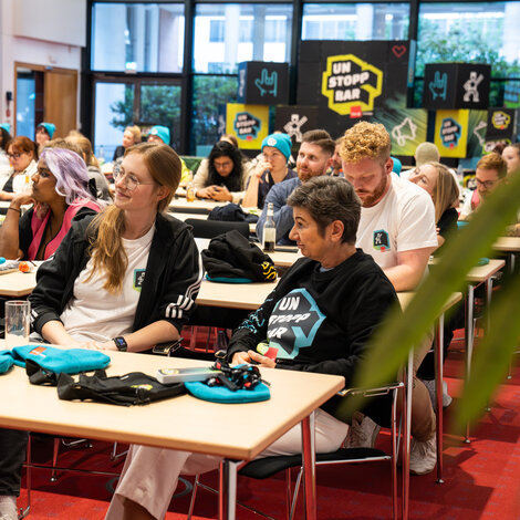 viele Menschen sitzen in einem Konferenzraum und schauen zu einer Bühne. Im Hintergrund sind Würfel mit UNSTOPPBAR-Design.